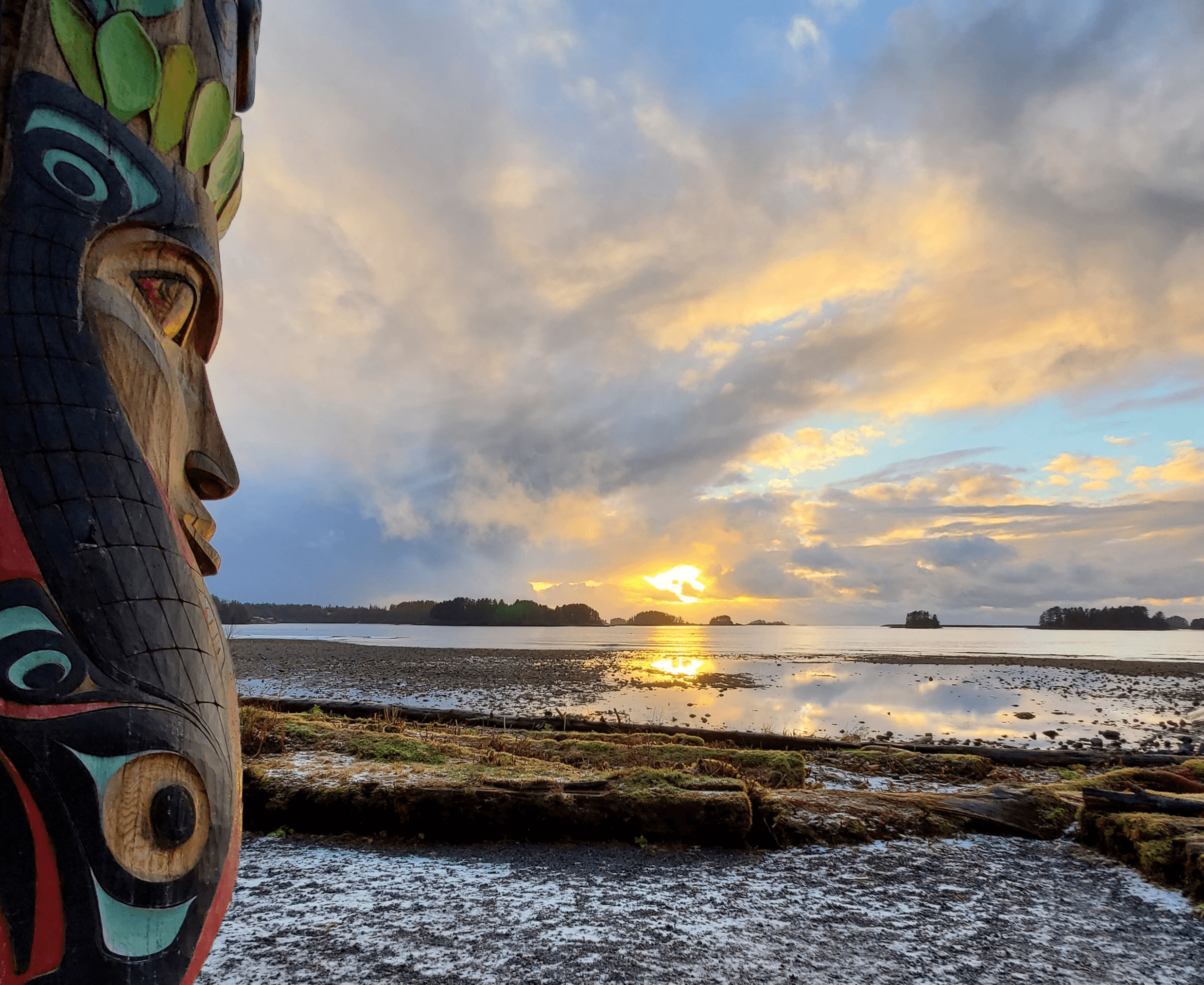 A totem pole in the foreground with a sunset over water in the background.